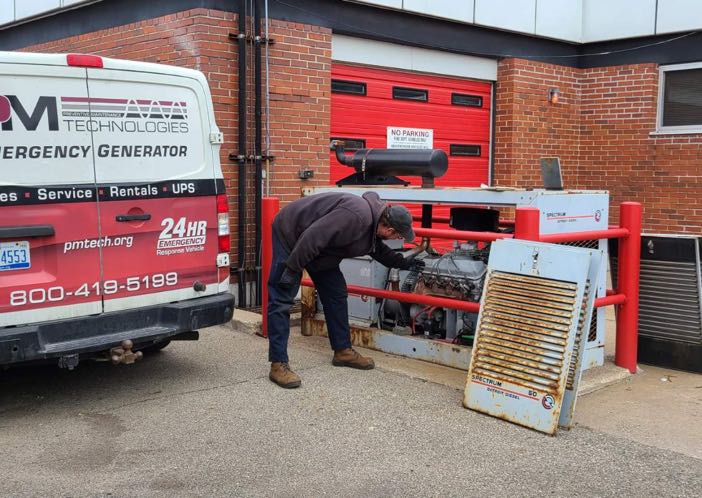 A PM Technologies branded vehicle is parked outside of a building. A technician is standing behind the vehicle and is bent over at the waist inspecting a commercial generator.